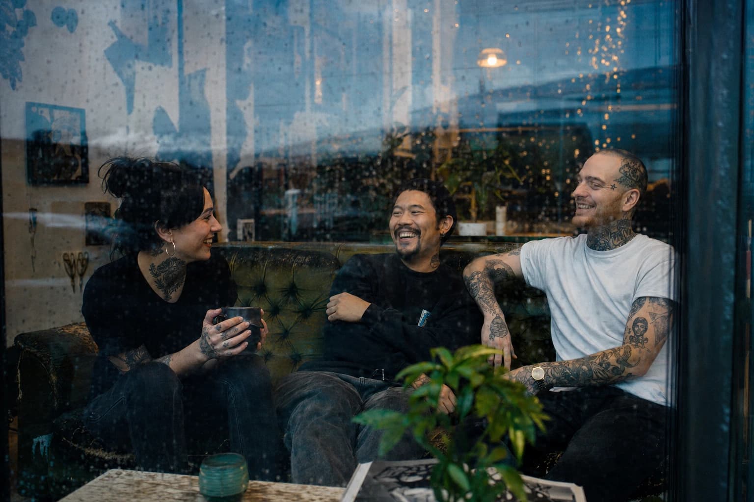 Tattoo shop crew laughing together through a rain-streaked window